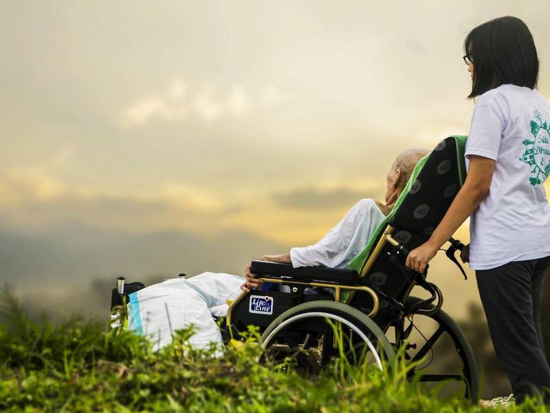 Mlajša oseba drži voziček z obnemoglo starejšo osebo in gledata sončni zahod